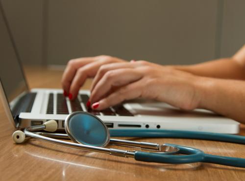Person typing on a laptop and stethoscope next to them
