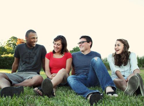 Group of young people sitting on grass. 4 people, left to right (man in shirt, woman with glasses, man with glasses, woman smiling)
