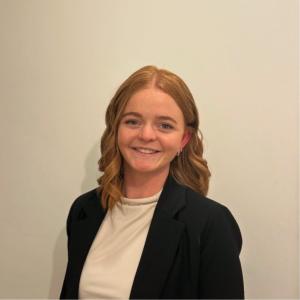 Rhiannon has ginger hair and is wearing a white top and black blazer