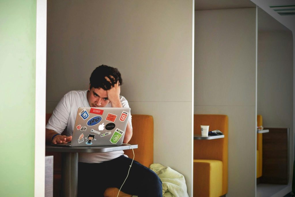 A young man sits in a small booth, leaning forward with one hand on his head while concentrating on a laptop covered in tech-related stickers, suggesting focus and possible stress while working. 